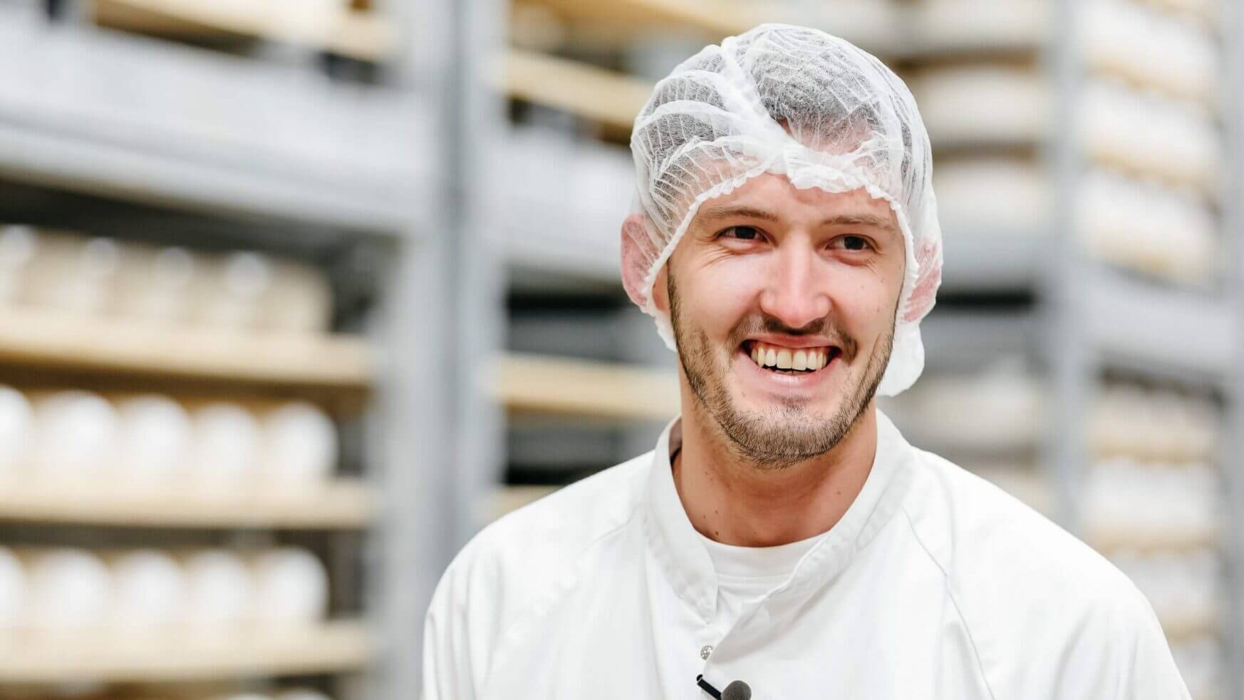 Lachende man in een wit pak en haarnetje staat in een opslagruimte met planken vol ronde voorwerpen.