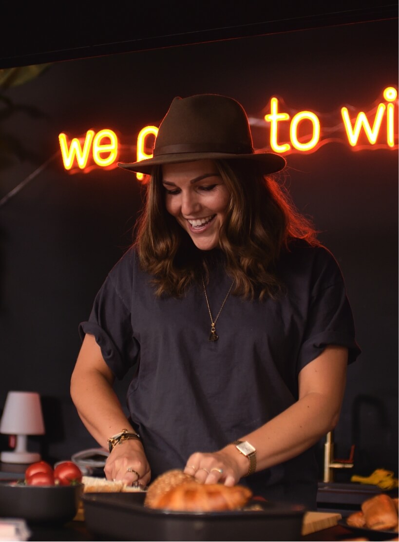 Een vrouw met een hoed snijdt brood in een keuken, verlicht door een neonreclame op de achtergrond.