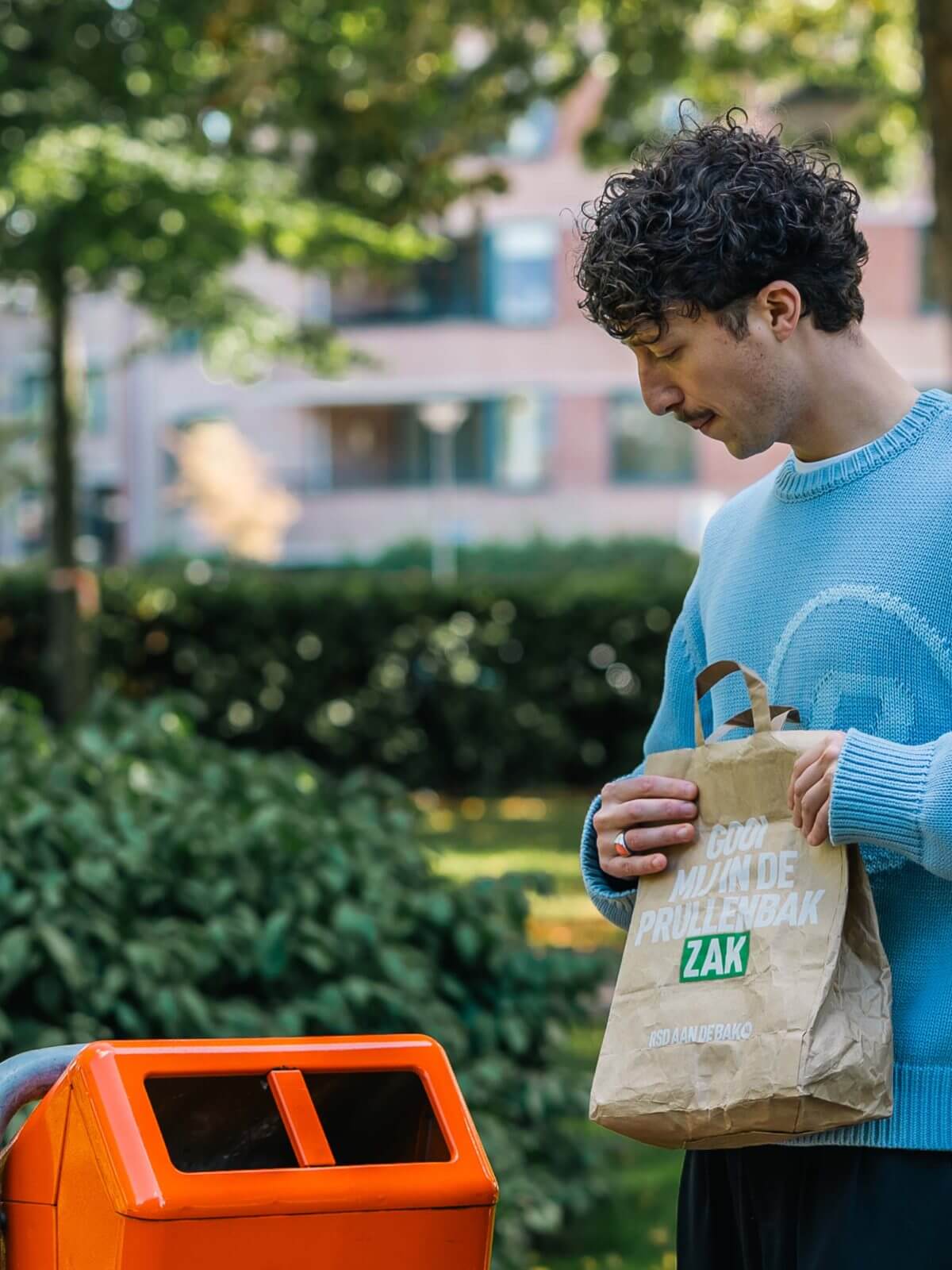Man in blauwe trui gooit papieren zak in oranje recyclebak buiten-RSD aan de bak!.