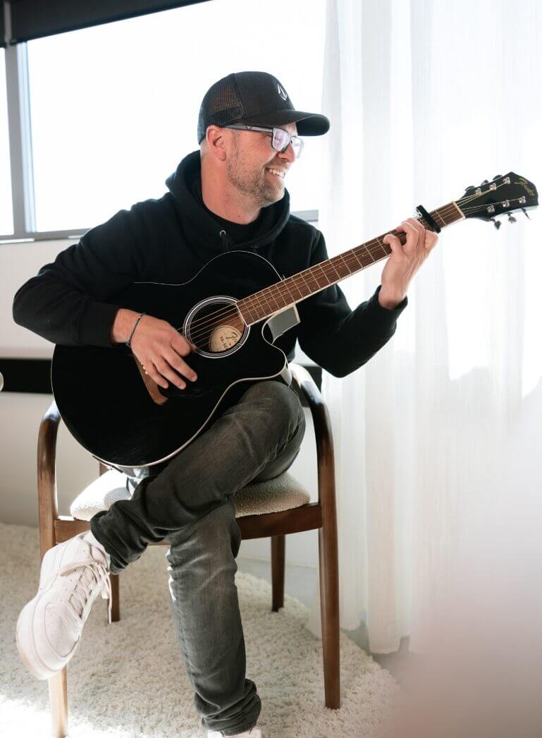 Iemand met een pet en een bril bespeelt een akoestische gitaar in een felverlichte kamer.