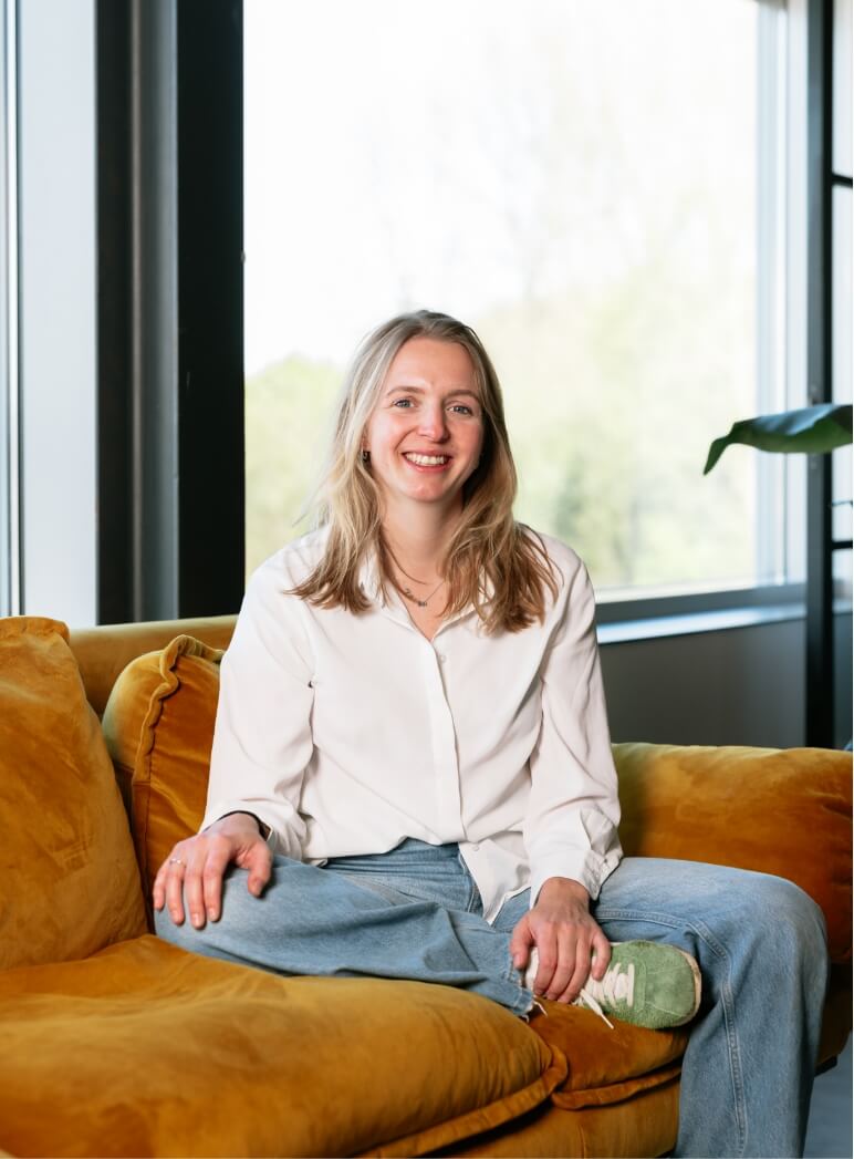 Met gekruiste benen op een mosterdgele bank, glimlachend in een wit shirt en spijkerbroek bij een groot raam.