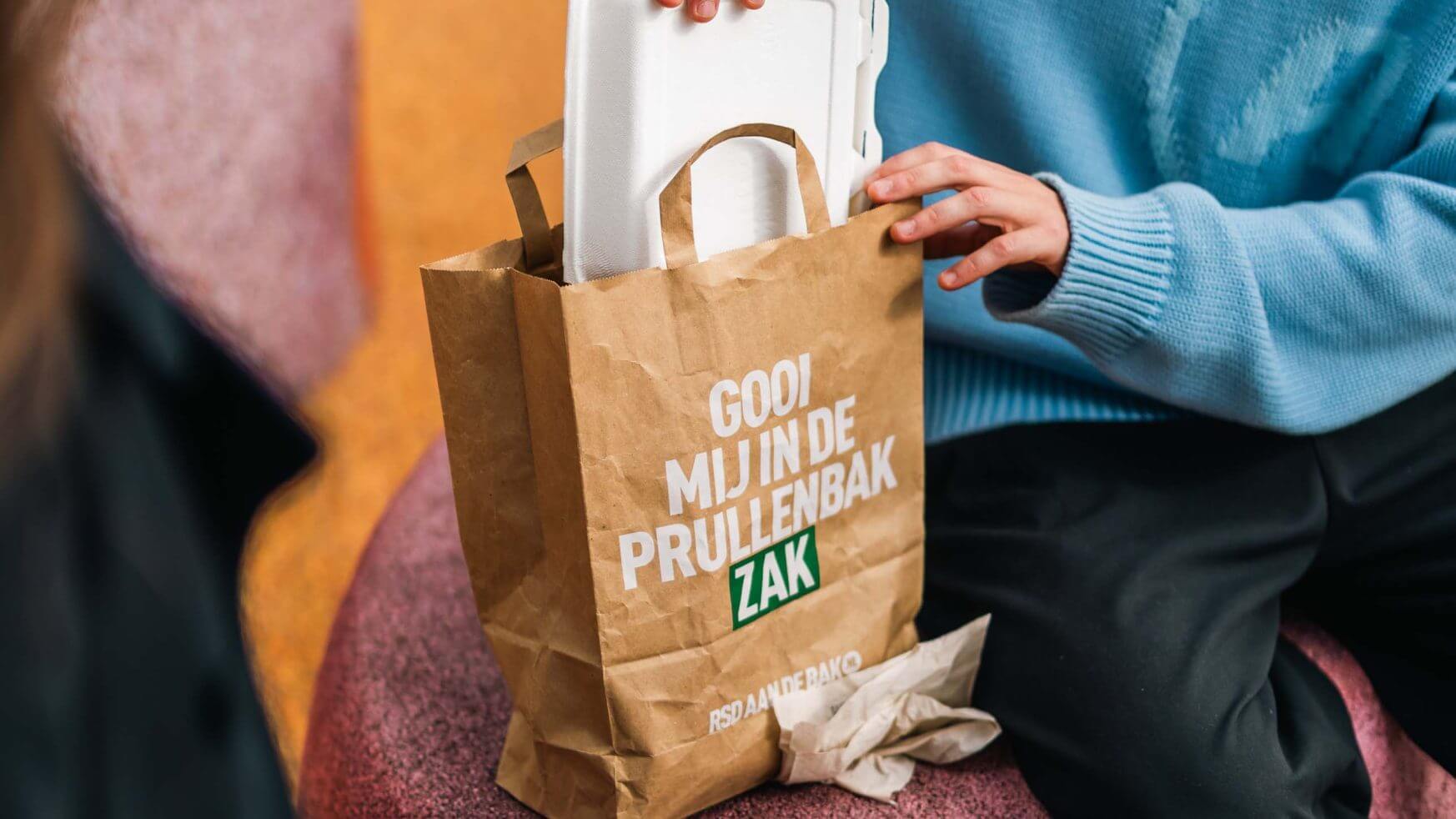 Persoon plaatst een witte voedselcontainer in een bruine papieren zak met het label "RSD aan de bak!" voor recycling of afval.