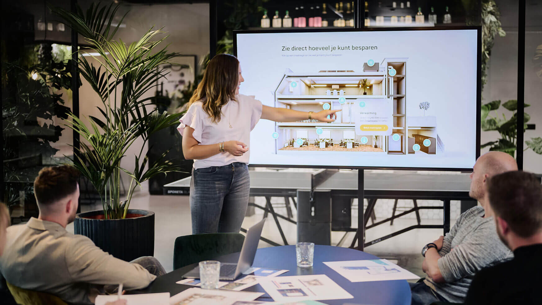 Een vrouw laat op het scherm een plattegrond zien aan drie collega's in een modern kantoor.