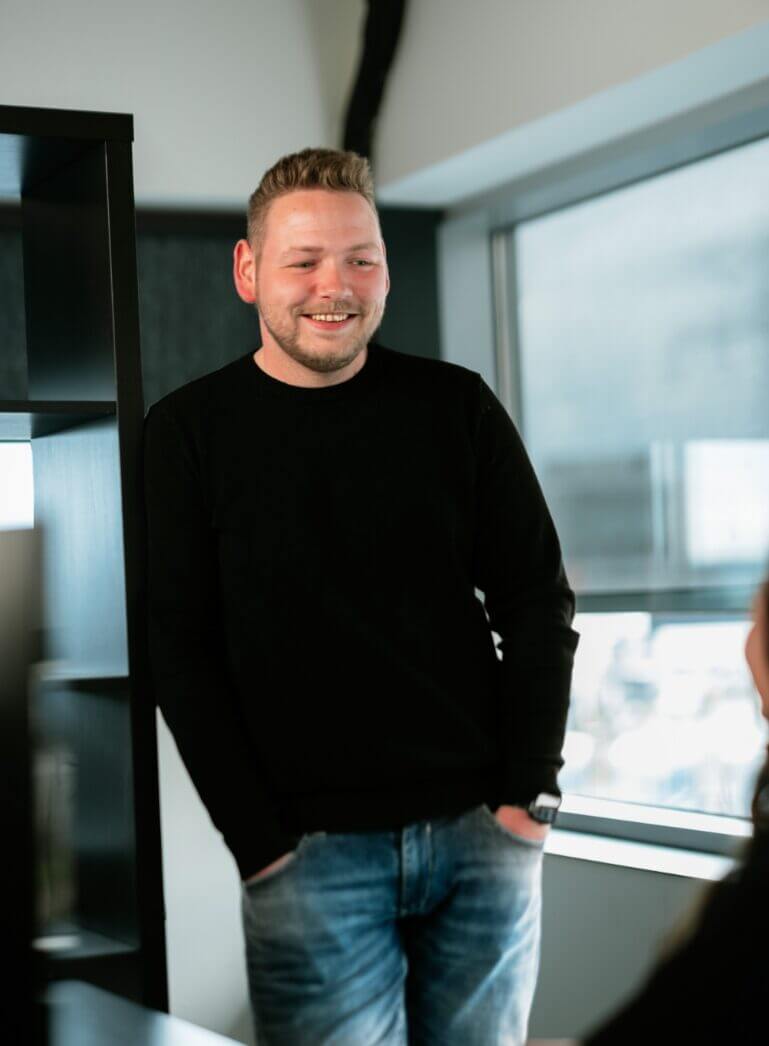 Een persoon in een zwarte trui en spijkerbroek staat lachend bij het raam in een lichte kamer.