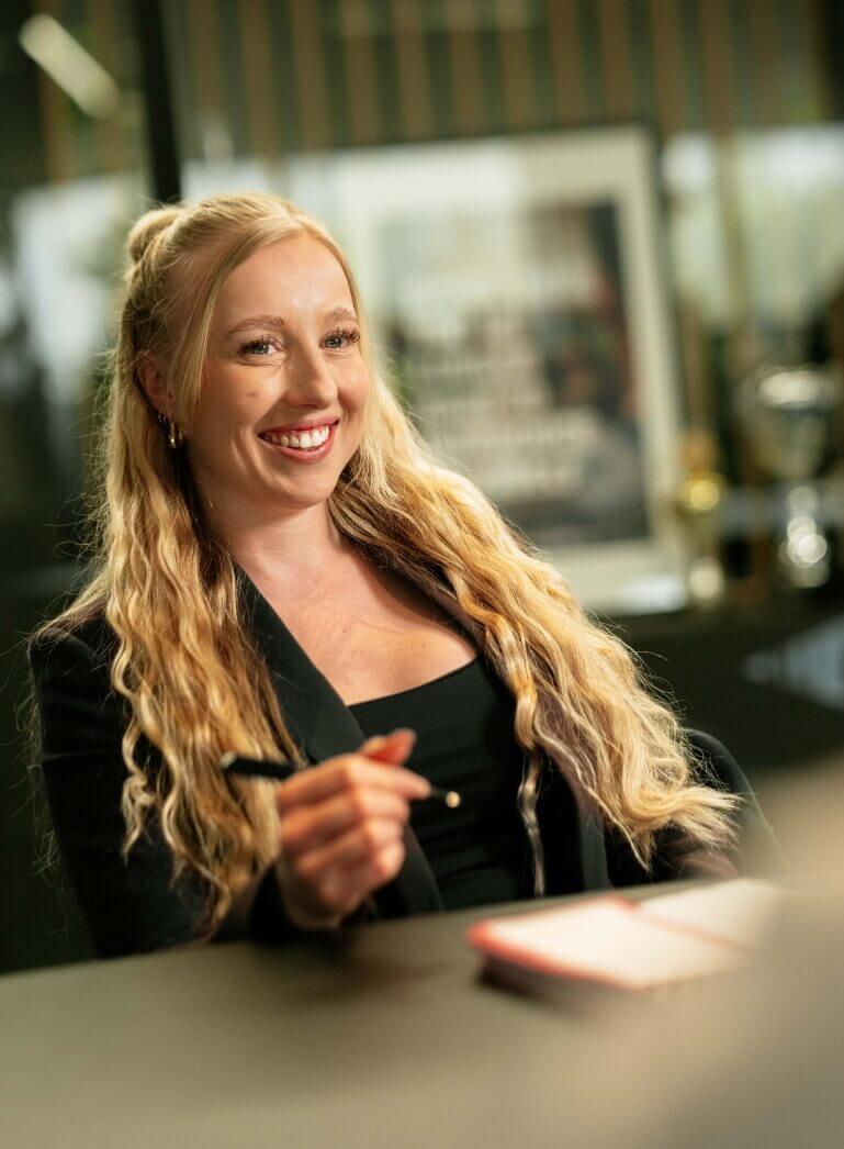 De blonde vrouw lacht en houdt een pen vast aan tafel.