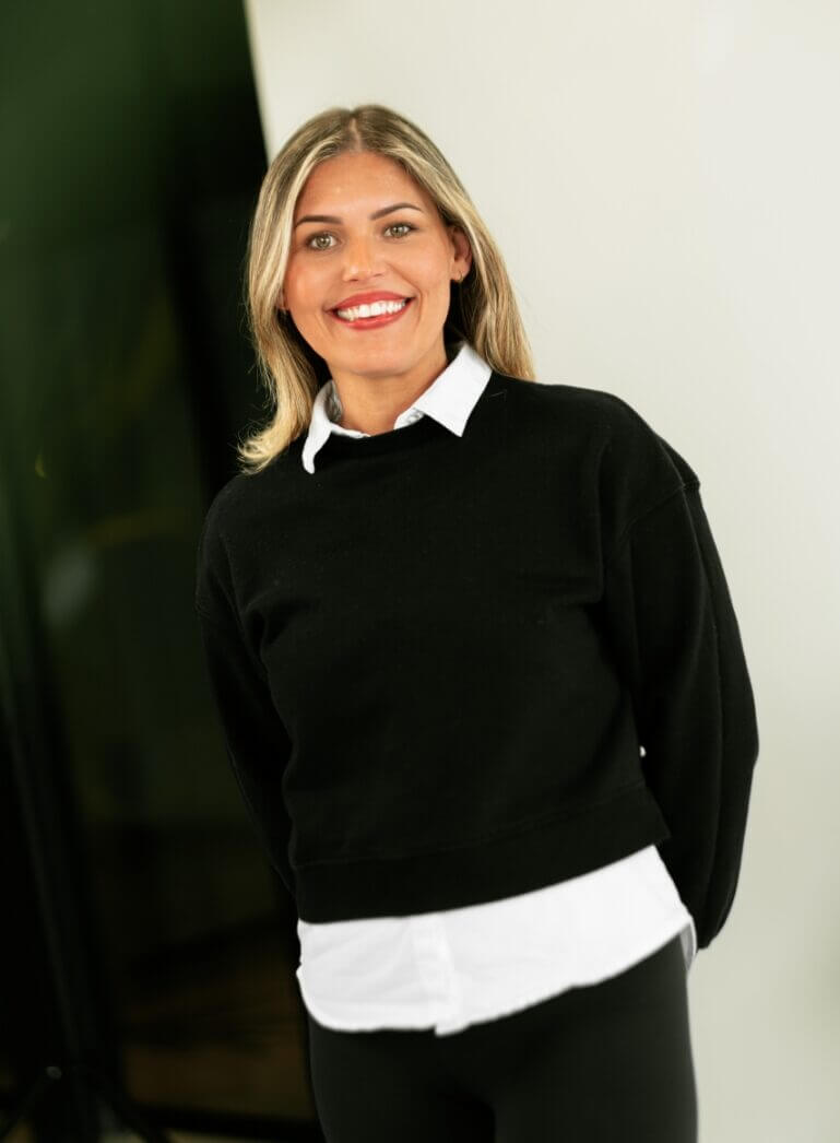 Een persoon glimlacht, draagt een zwarte trui over een wit overhemd tegen een effen achtergrond.