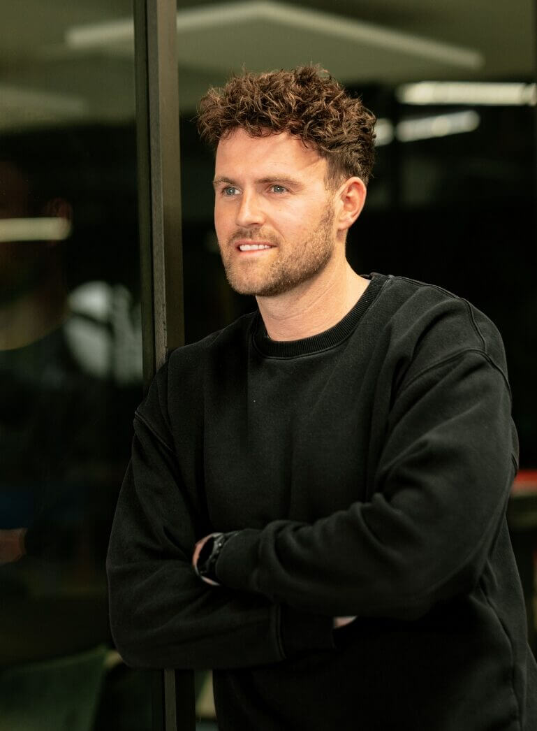 Een man met krullend haar en een baard in een zwarte trui staat lachend met zijn armen over elkaar.