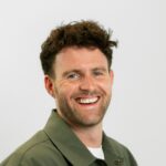 Man met krullend haar in een groen shirt, lachend tegen een witte achtergrond.