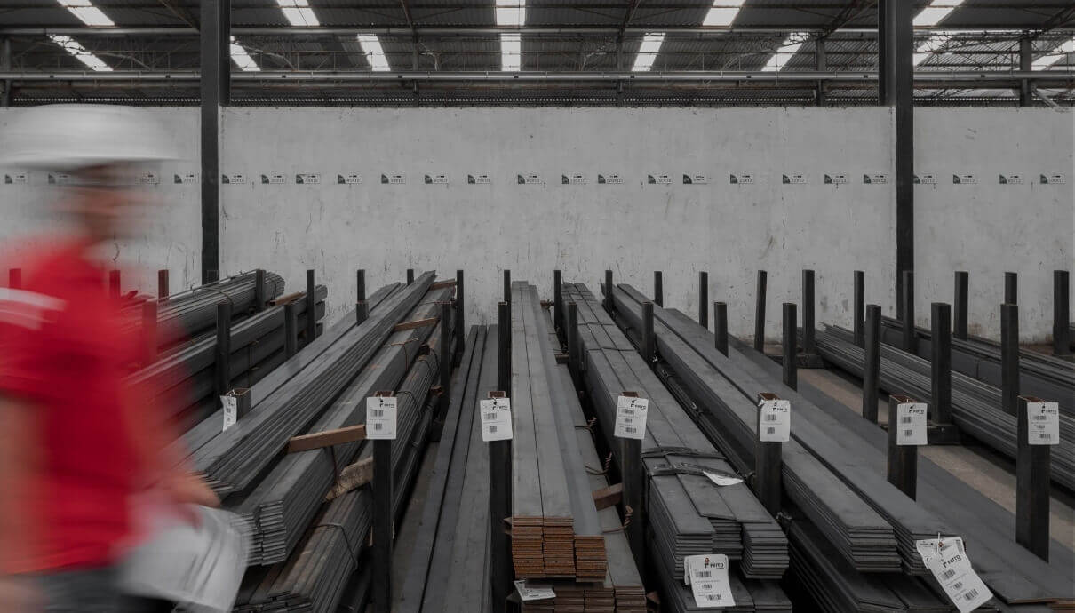 Een persoon in een rood shirt en een helm loopt langs opgestapelde metalen balken in een pakhuis.