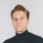 Lachende persoon met kort haar, zwarte coltrui, lichtgrijze achtergrond.