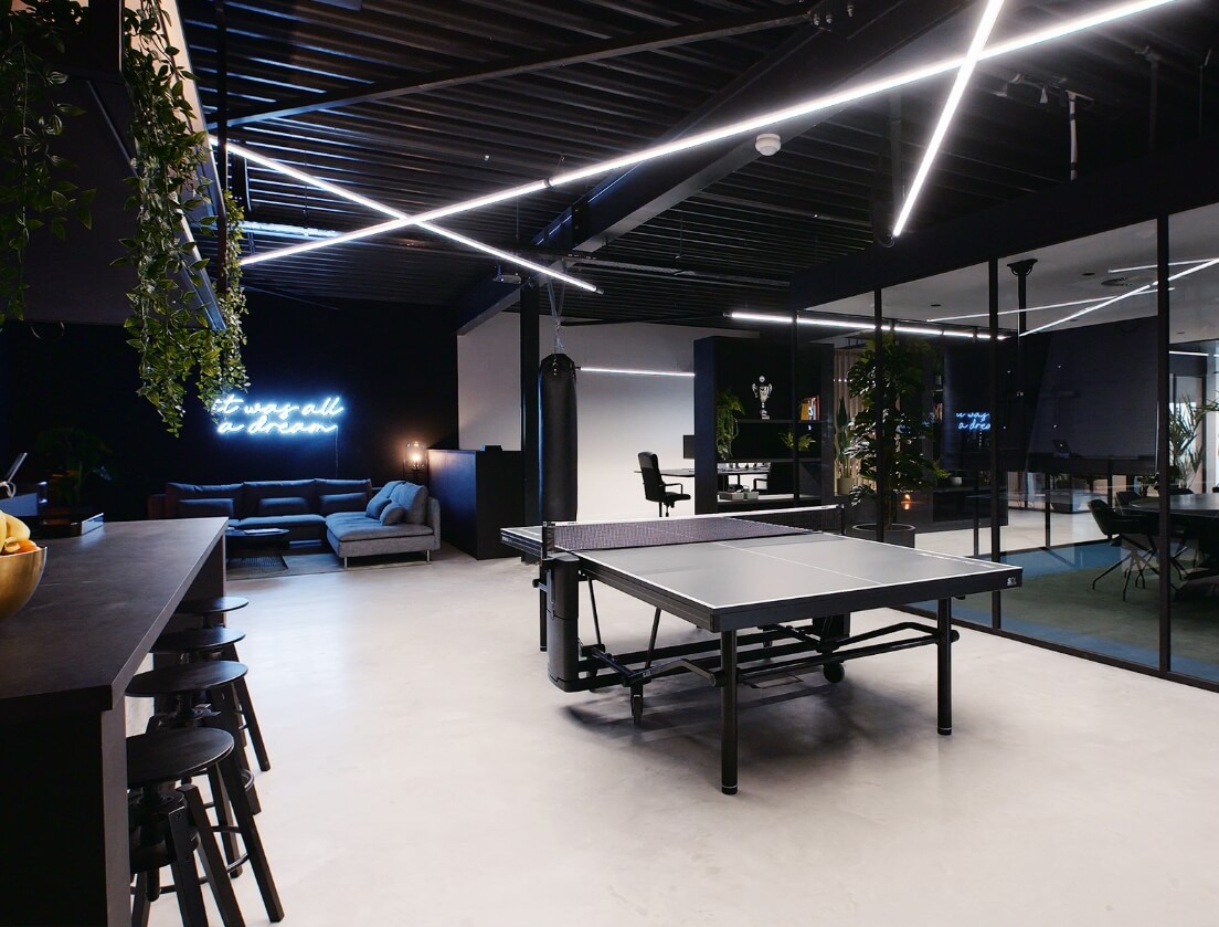Strakke kantoorlounge: pingpongtafel, comfortabele banken, vergaderruimte met glas, neonreclame tegen een donkere muur.