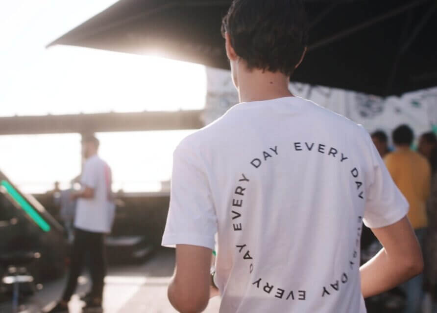 Een man in een wit EVERY DAY T-shirt loopt buiten, met zonlicht en wazige mensen op de achtergrond.