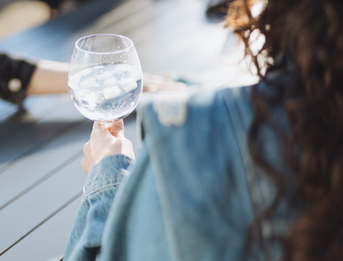 Persoon in spijkerbroek houdt ijswater vast aan een tafeltje buiten; een andere hand is zichtbaar op de tafel.