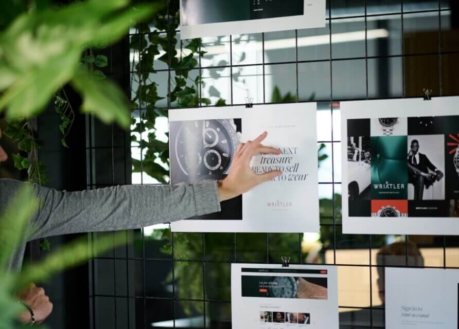 Een hand wijst naar designposters op een draadraster, omgeven door weelderige groene planten.
