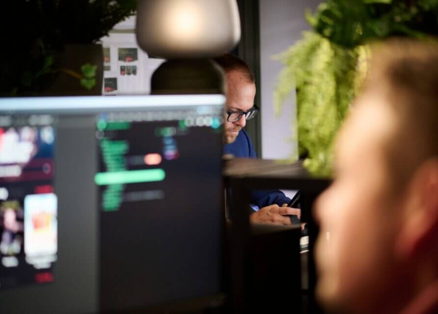 Programmeur achter computer in focus; persoon met planten vaag op de achtergrond.