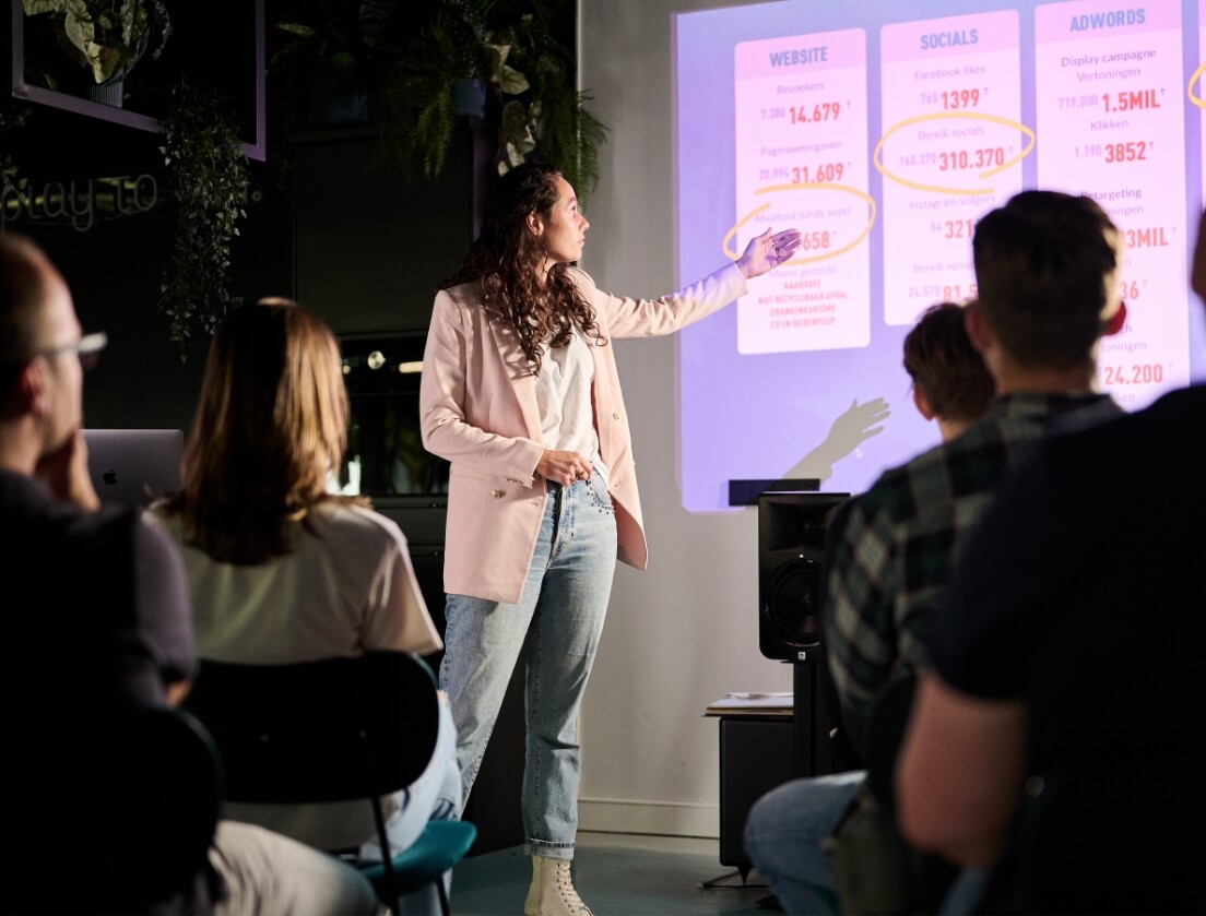 In een donkere kamer presenteert een vrouw in een wit jasje gegevens op het scherm aan een aandachtig publiek.