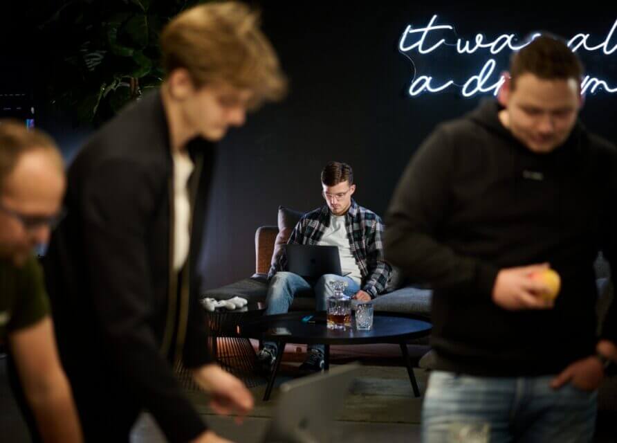 Modern kantoor: persoon achter laptop, anderen in de buurt; neonlicht op de muur.