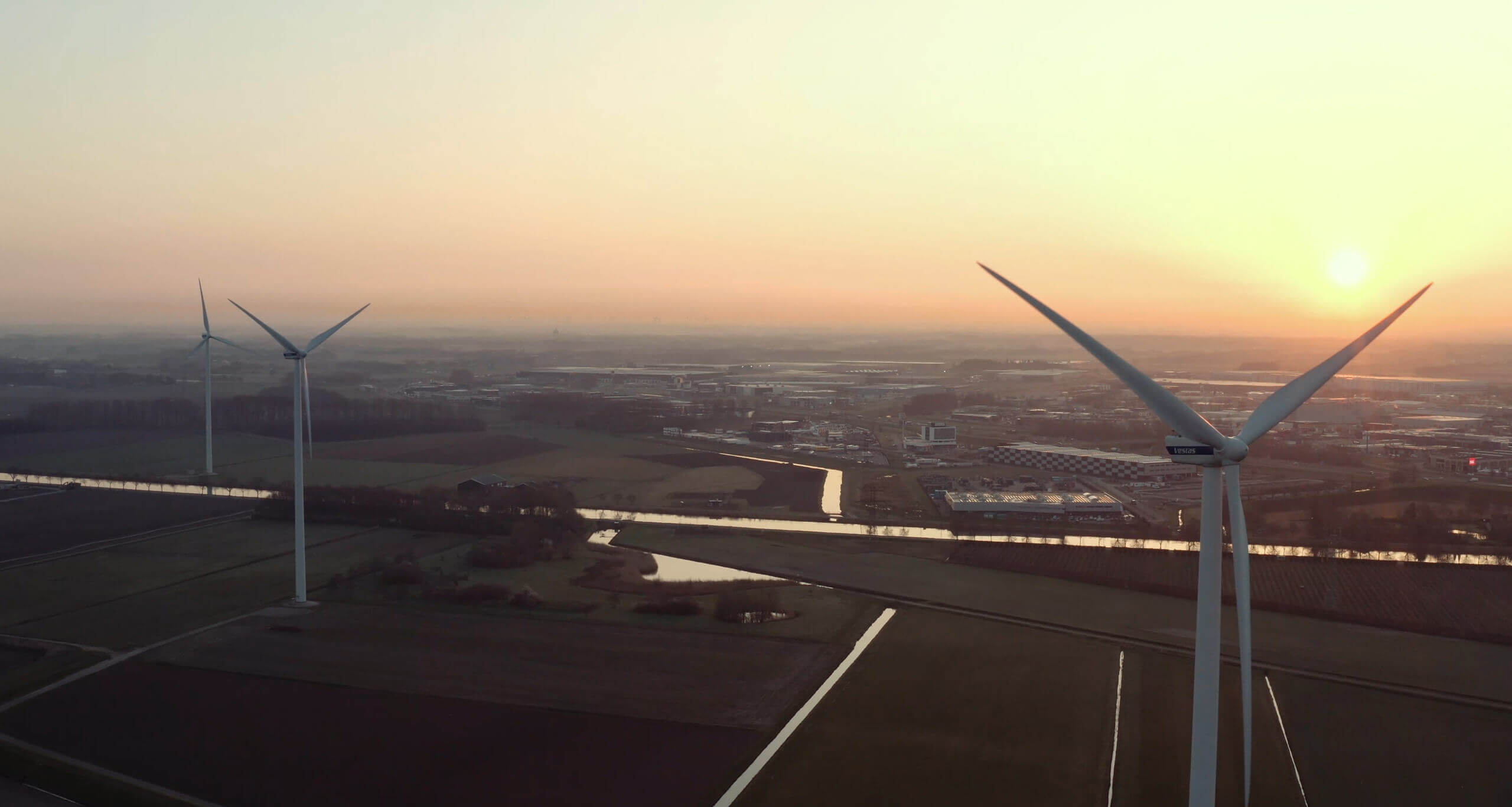 Luchtfoto van zonsondergang: windturbines staan verspreid over landelijke velden, een rivier slingert door het landschap.