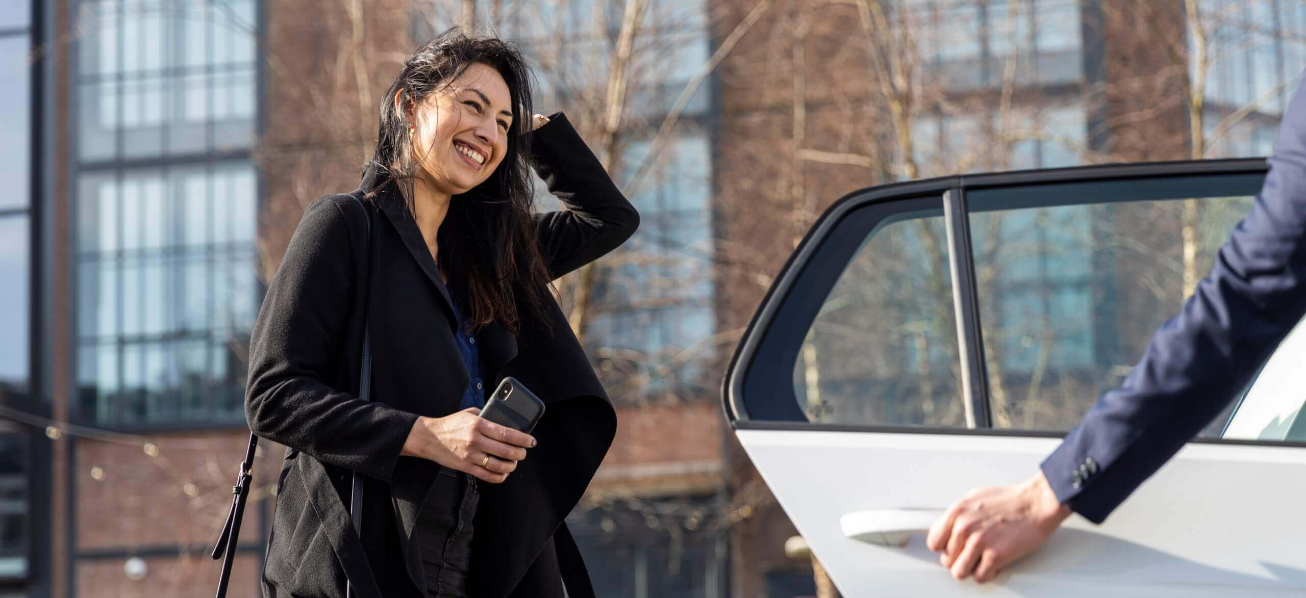 Een vrouw glimlacht, met de telefoon in haar hand, terwijl iemand anders naast haar de deur van een taxi opent.