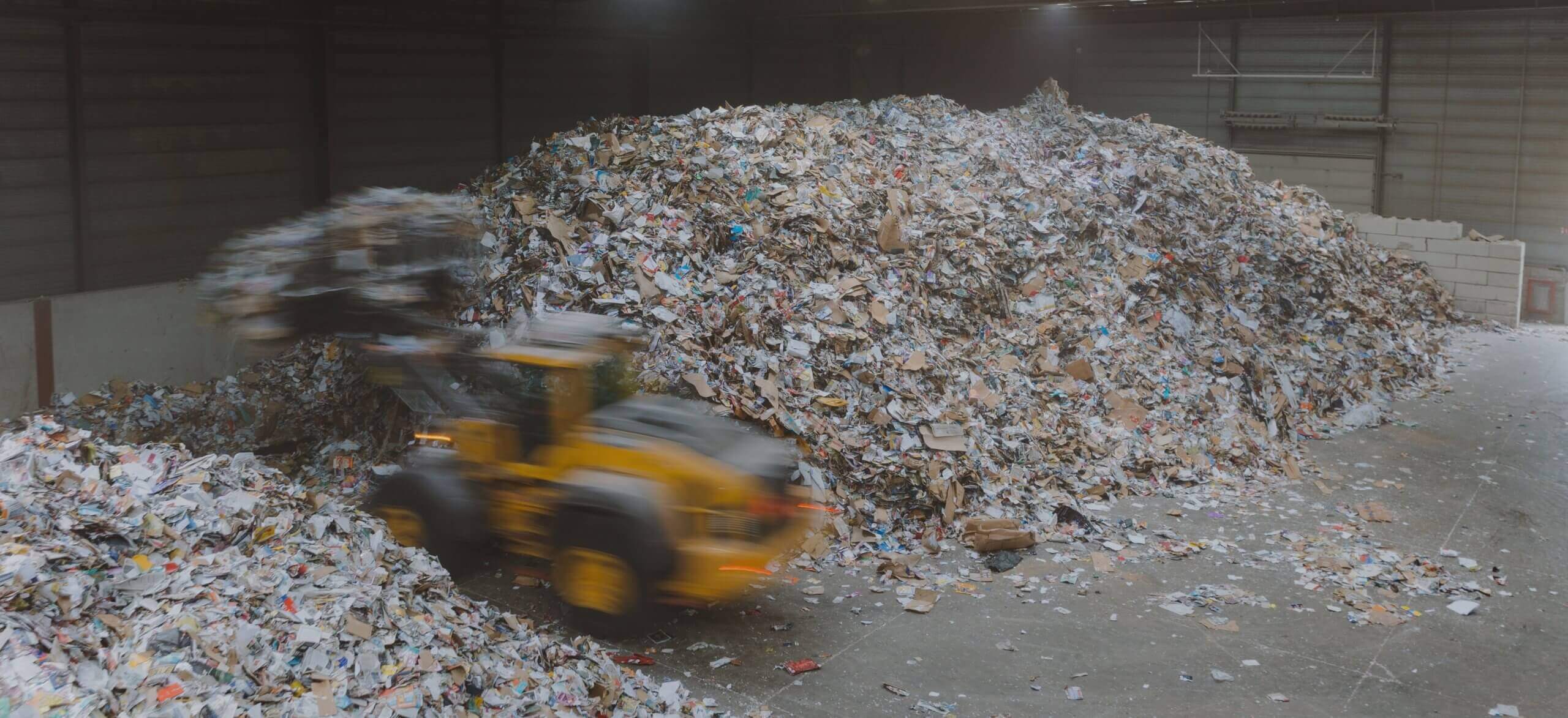 Een gele lader verplaatst papier en karton in een grote overdekte recyclingfaciliteit.