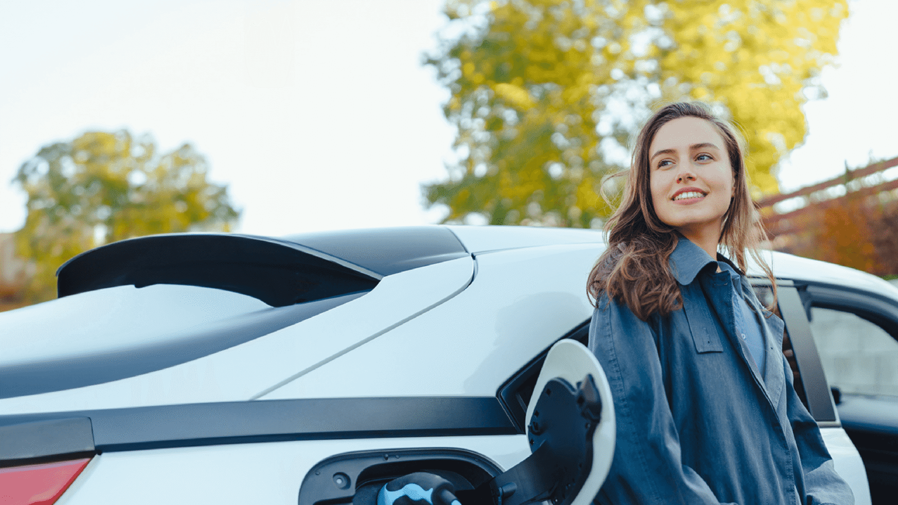 Een lachende vrouw staat bij een witte elektrische auto bij een laadstation, met bomen op de achtergrond.