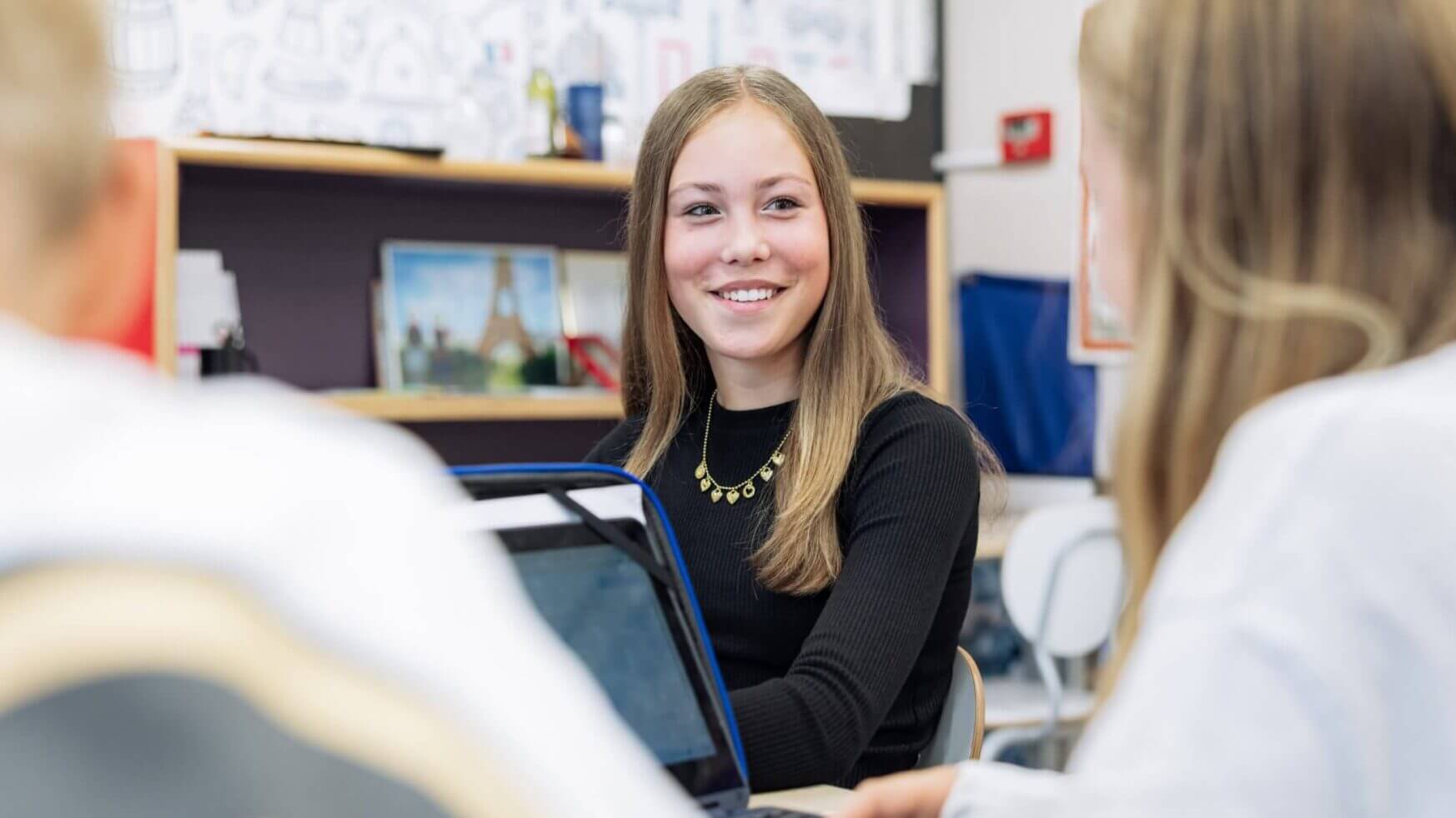 Een meisje praat lachend met haar klasgenoten, terwijl ze voor haar in de klas een laptop openklapt.
