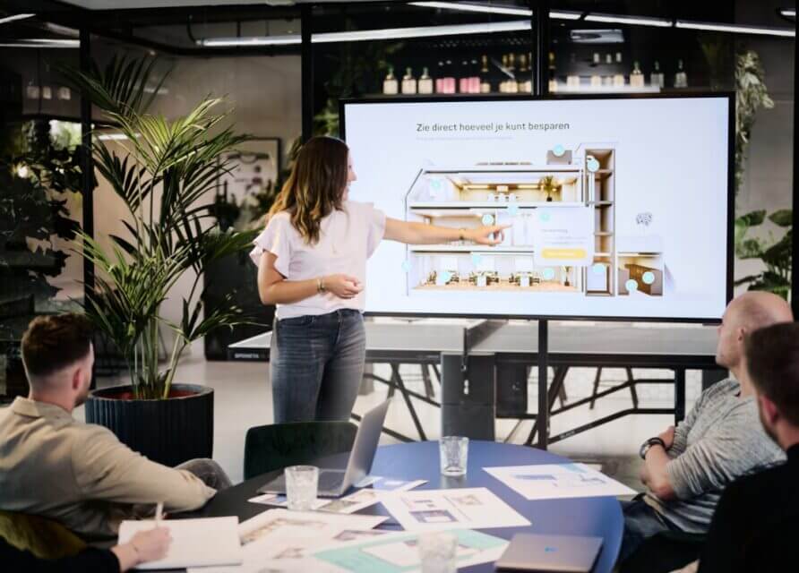 Een vrouw presenteert haar project op een scherm aan drie collega's in een modern kantoor.