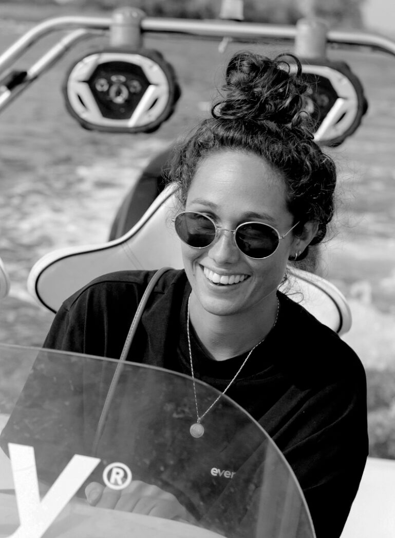 Een vrouw met een knotje en een zonnebril bestuurt een boot en lacht op het water.