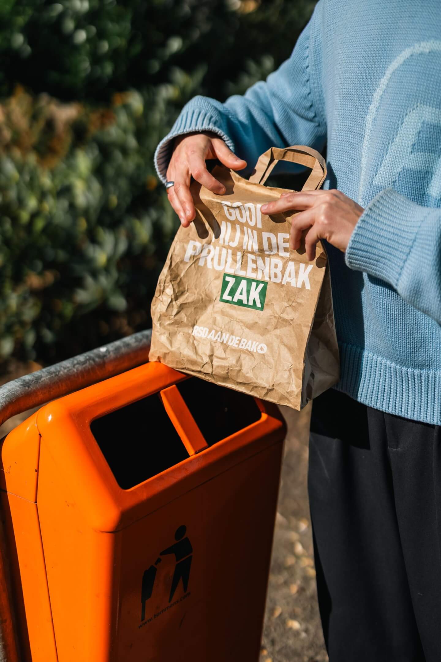Persoon met bruine papieren zak bij oranje prullenbak buiten. RSD aan de bak! roept op tot verantwoorde afvalverwerking.