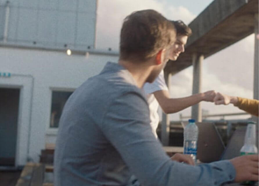 Drie op een dak: twee fist bumps, een derde in blauwe horloges. Laptops en drankjes verspreid over de tafel.
