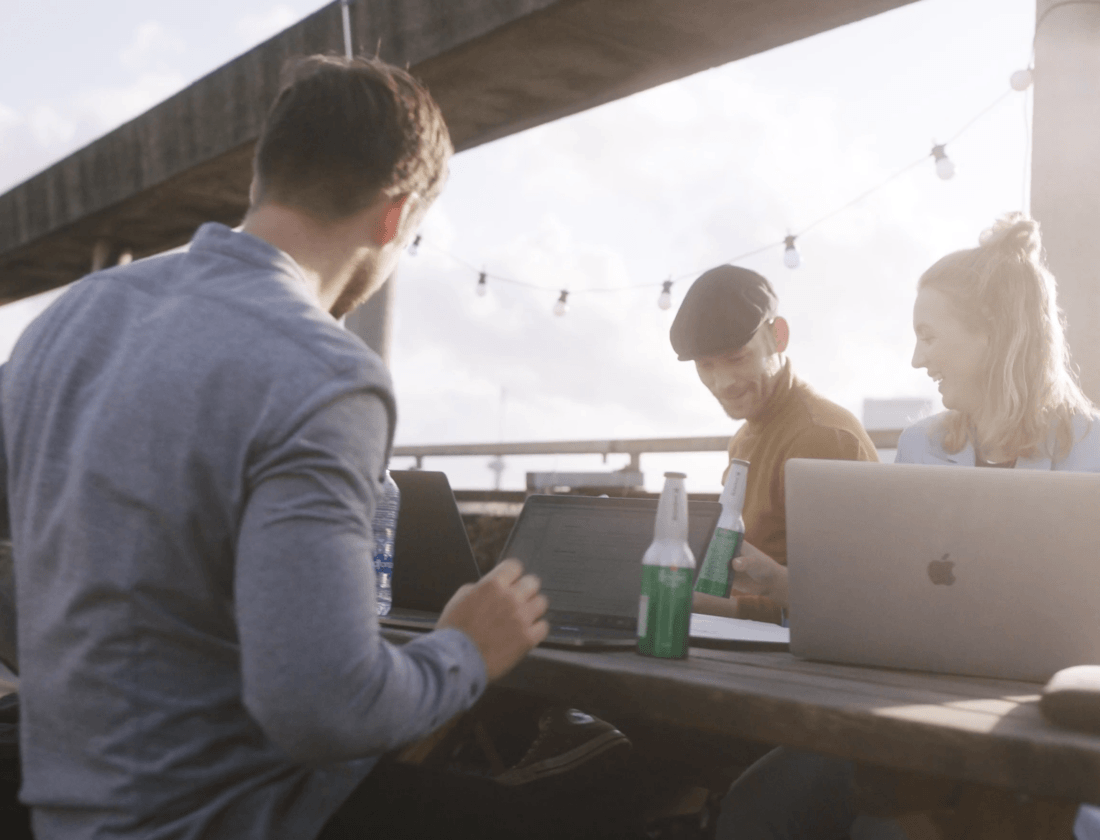 Drie mensen kletsen en lachen rond hun laptops aan een tafeltje buiten, met daglicht en lichtslingers erboven.