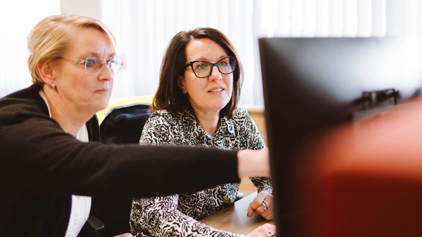 Twee geconcentreerde vrouwen zitten aan een bureau, waarvan er één naar het computerscherm wijst.