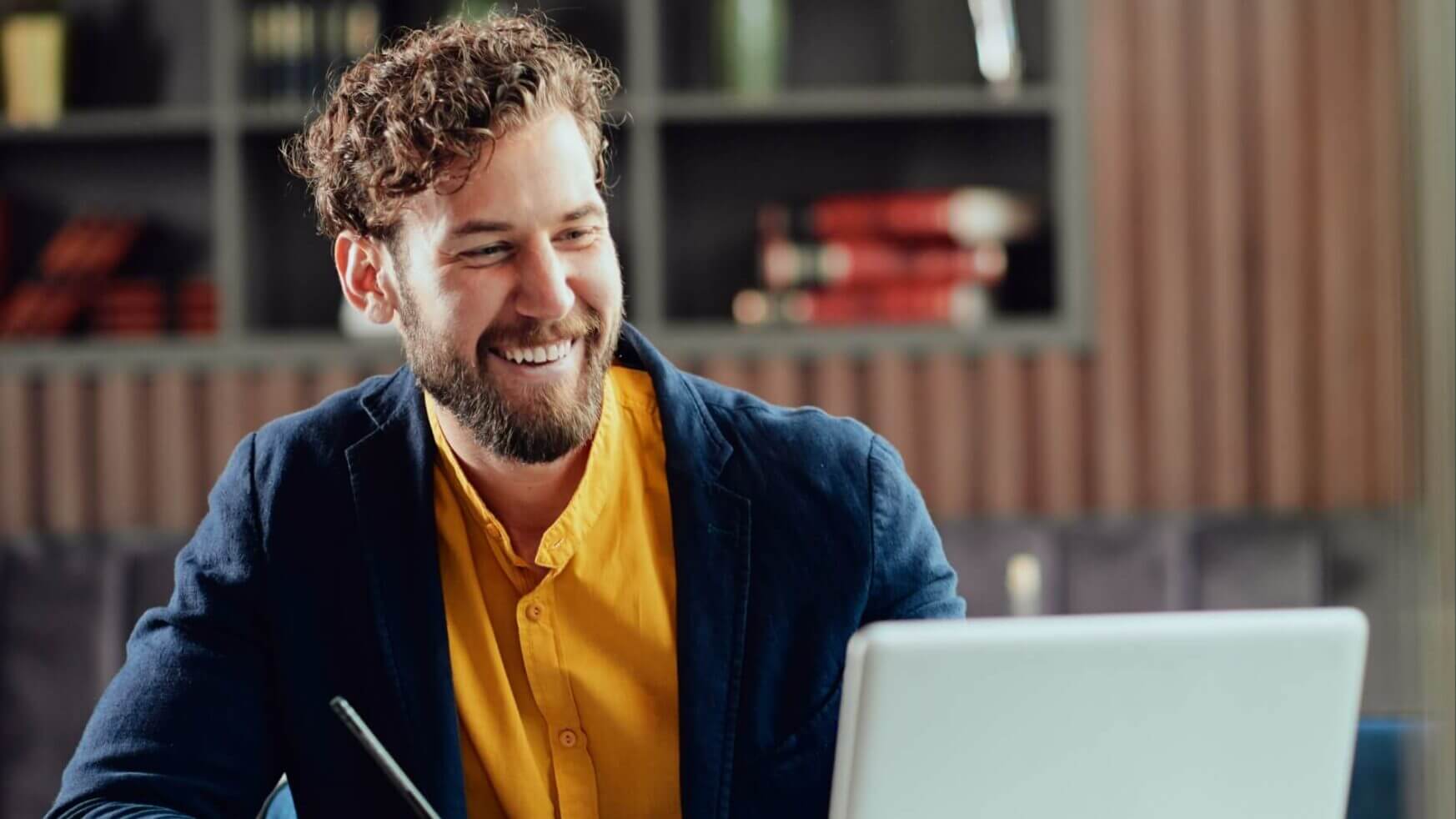 Lachende man in een geel overhemd en een blauw jasje typt op zijn laptop en schrijft met een pen in een modern kantoor.