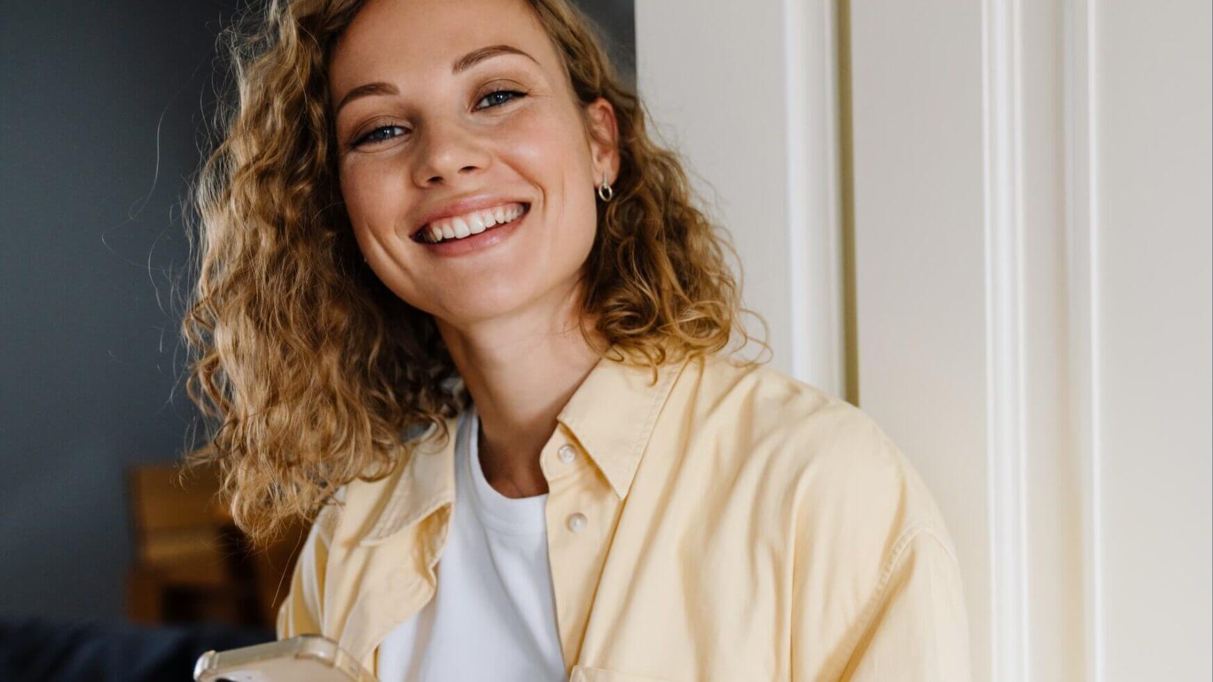 Een persoon met krullend haar in een geel shirt houdt een telefoon vast en lacht bij een witte deur binnenshuis.
