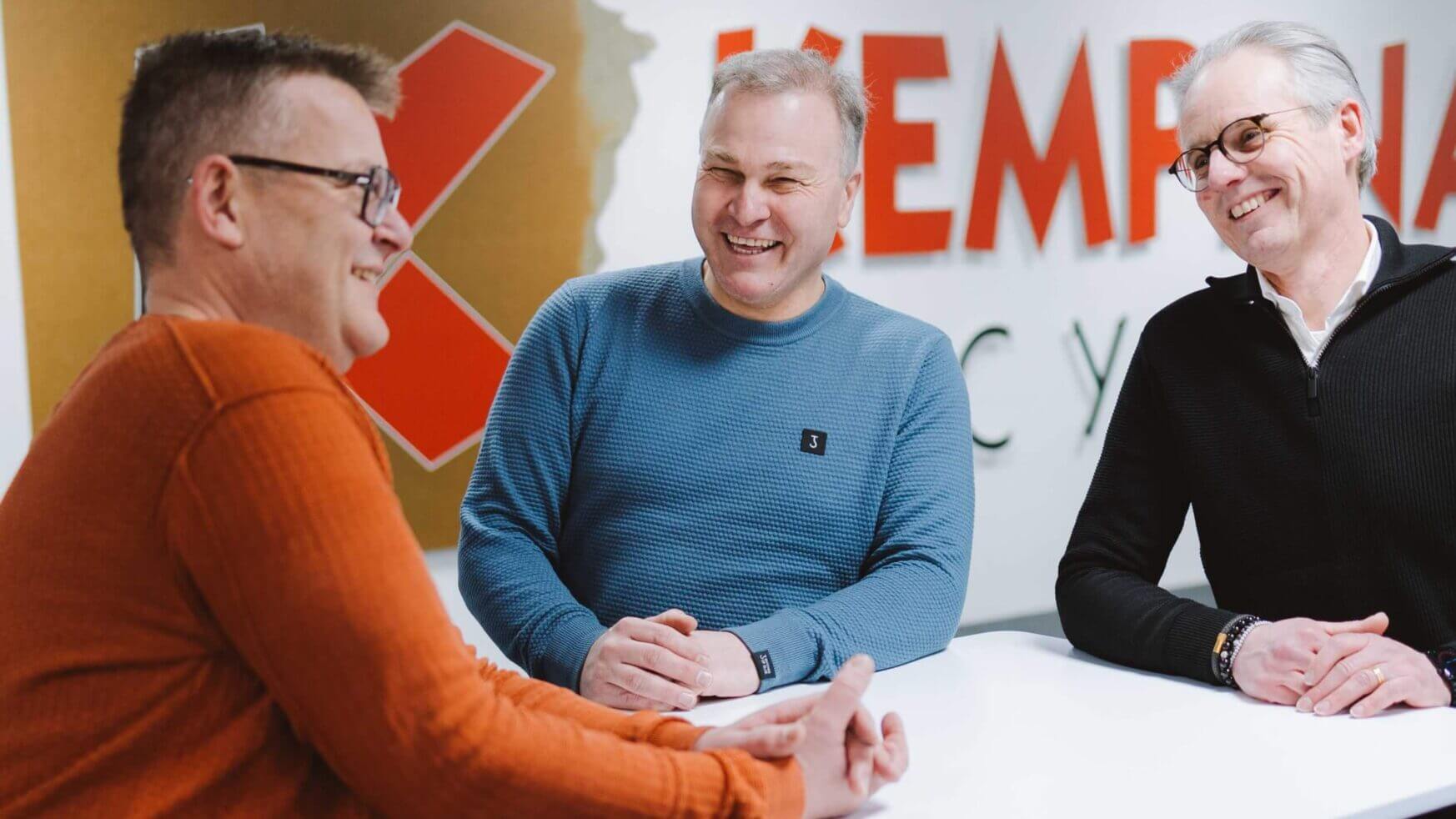 Drie mannen kletsen en lachen rond een witte tafel in een strak, modern kantoor.