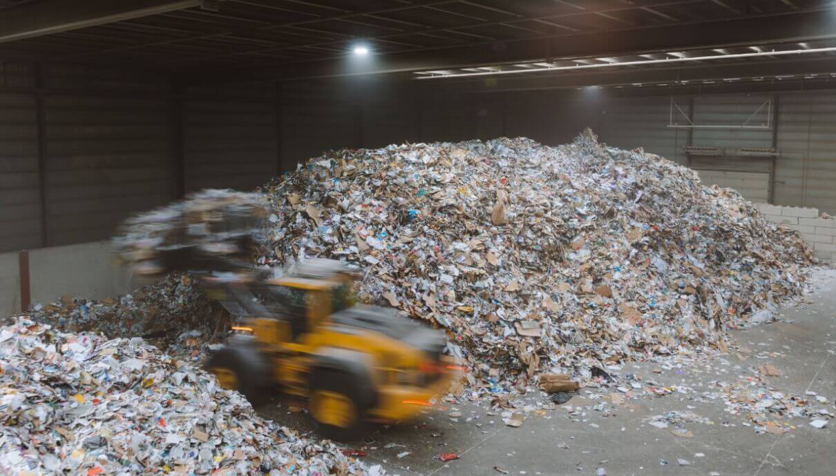 Een gele bulldozer rijdt langs stapels papierafval in een slecht verlichte recyclingfabriek.