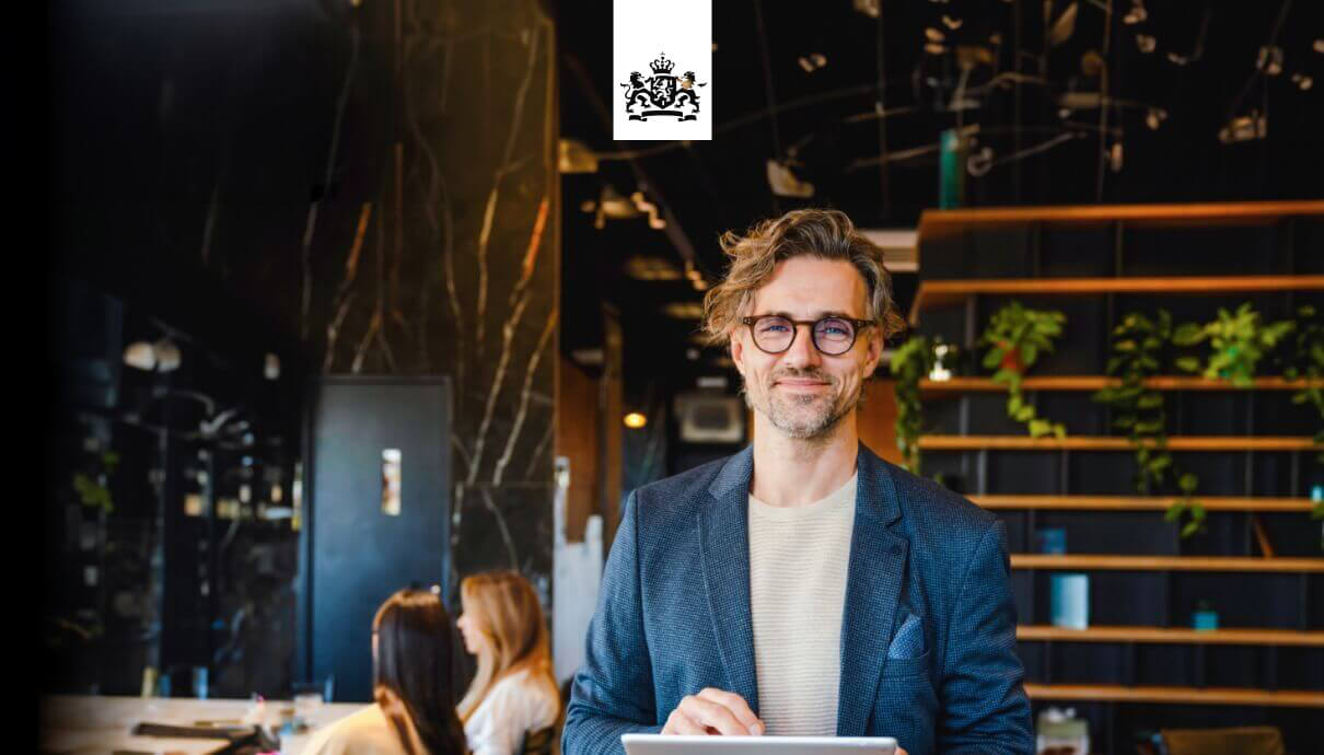 Een glimlachende man met een bril en een tablet in een modern kantoor, met op de achtergrond planten en collega’s die aan het werk zijn.