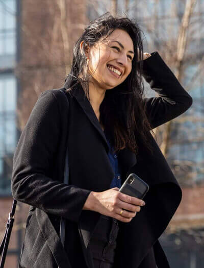 Een lachende vrouw in een zwarte jas houdt haar telefoon vast en wacht op een taxi op een zonnige dag.