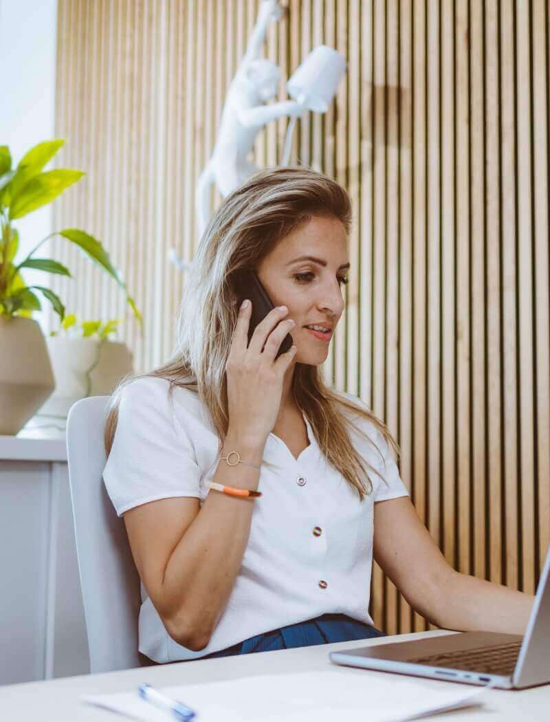 Vrouw aan een bureau, telefoon in de hand, laptop open, houten achtergrond en potplanten sieren het tafereel.