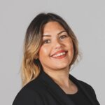 Vrouw met lang haar, glimlachend in een zwart jasje tegen een grijze achtergrond.