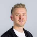 Lachend, kort blond haar, zwart jasje, wit overhemd, lichtgrijze achtergrond.