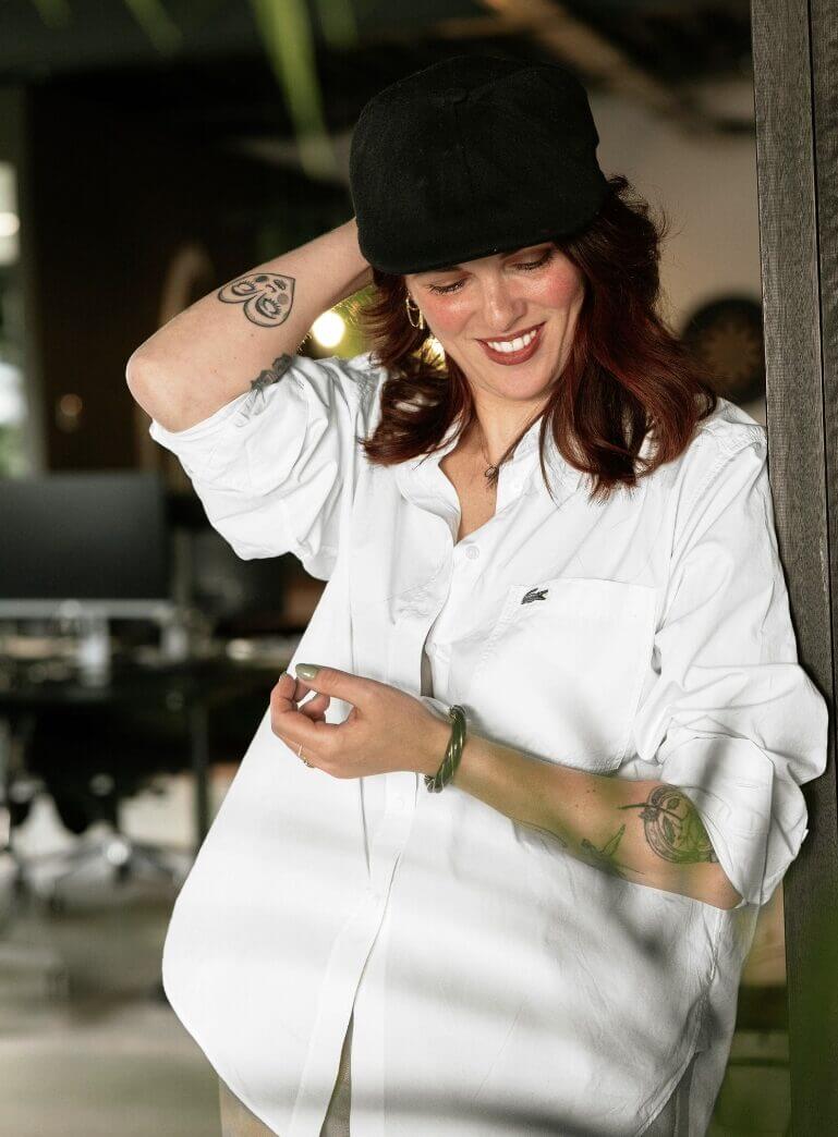 Lachende persoon in een wit overhemd en een zwarte pet, die één arm omhoog houdt naast een houten balk binnenshuis.