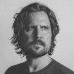 Zwart-witportret: man met schouderlang haar, een baard, die opzij kijkt.
