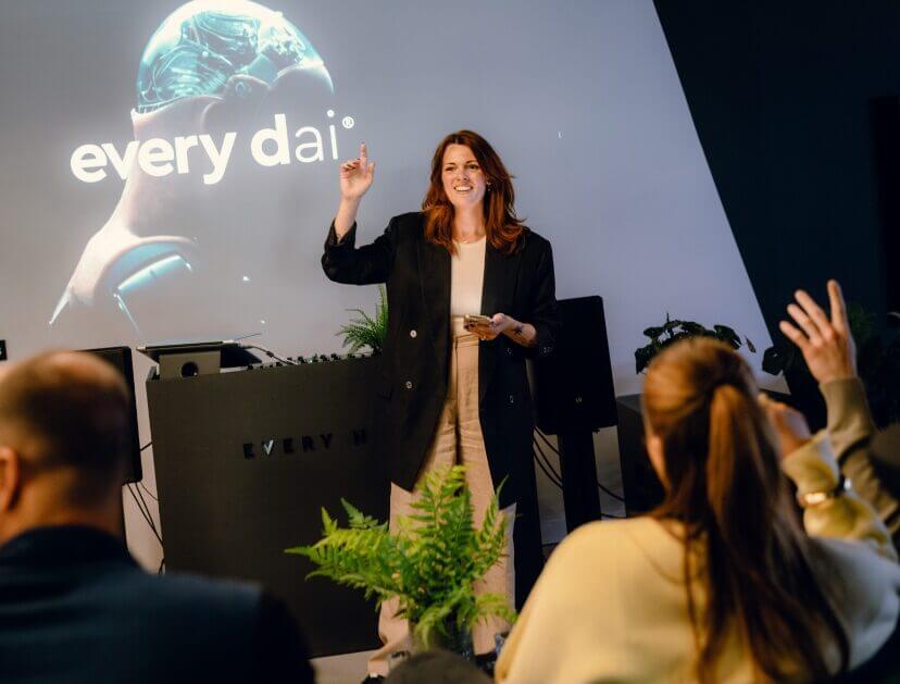 Een vrouw geeft een presentatie aan het publiek. Ze gebruikt een scherm waarop 'elke dag' en een afbeelding van een robot te zien zijn.