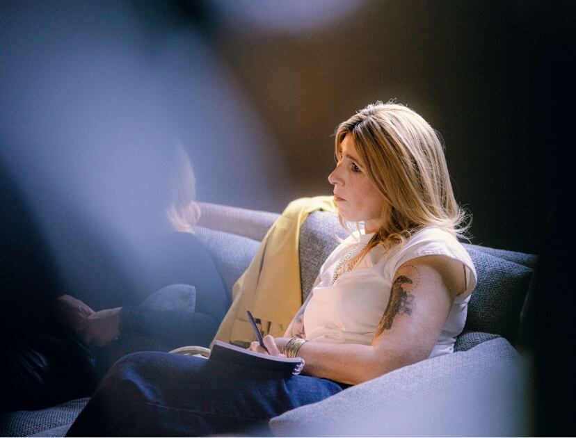 Een vrouw met lang haar zit op een bank, notitieboekje en pen in de hand, in gedachten verzonken.