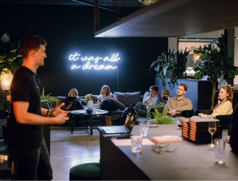 Een man spreekt een groep mensen toe in een lounge. Achter hem staat een neonbord met de tekst: "Het was allemaal een droom.