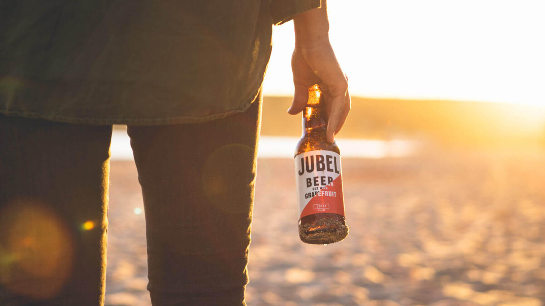 Terwijl de zon ondergaat en het strand goud kleurt, geniet iemand van een verfrissend Jubel-biertje.