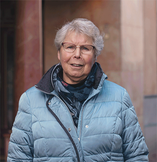 Een oudere vrouw, lachend met een bril en een blauw jasje, staat buiten tegen een wazige architectuurachtergrond.