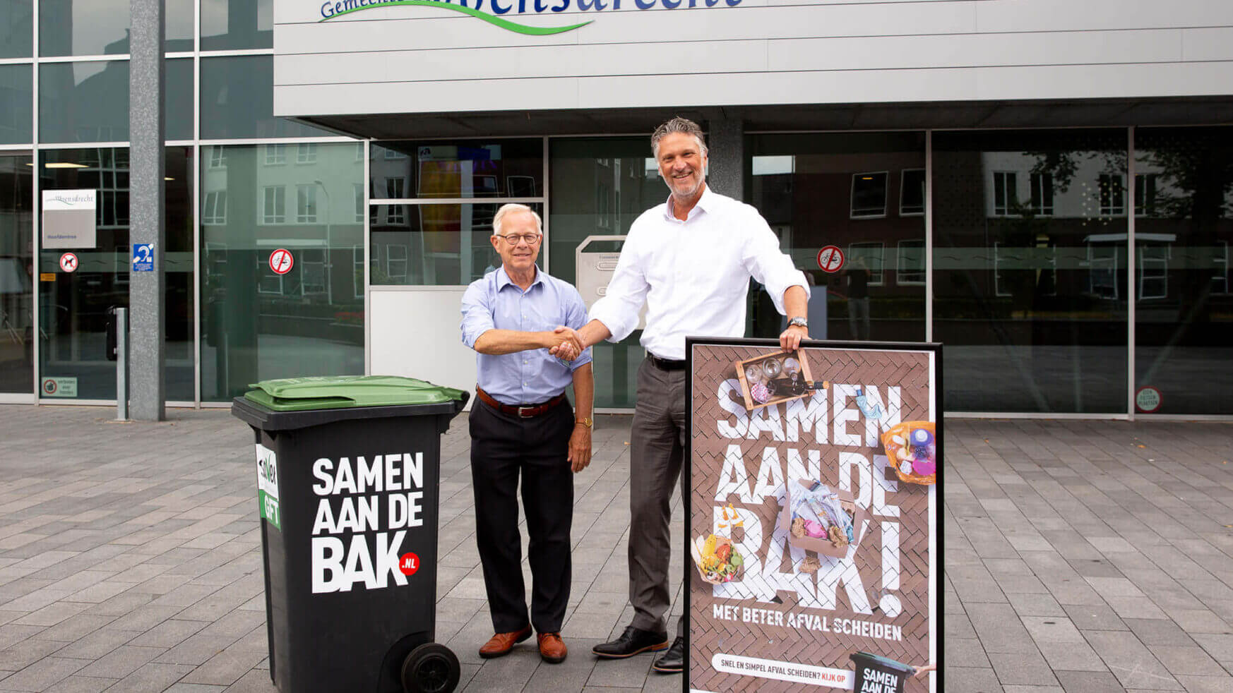 Twee mannen schudden elkaar de hand bij een prullenbak en "Samen Aan De Bak!" affiche in Woensdrecht.