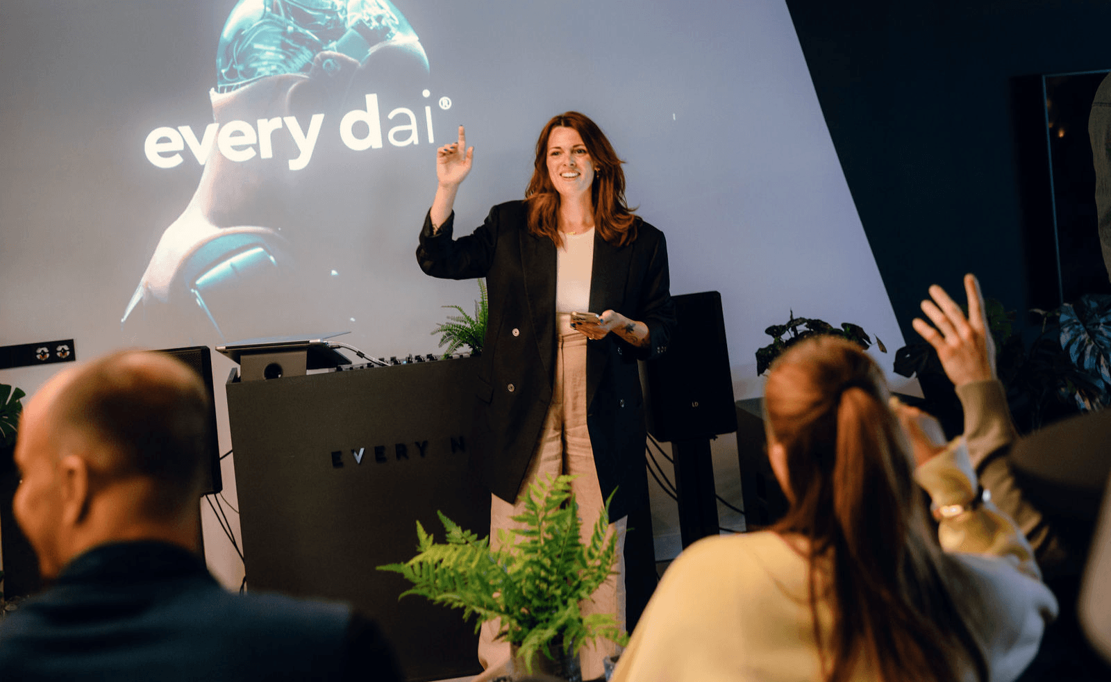 Een vrouw geeft een presentatie aan een publiek, met achter haar op het scherm 'elke dag' strakke, futuristische graphics.